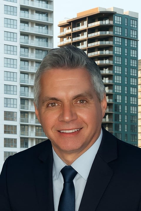 Professional headshot of middle-aged man in dark suit and tie, smiling at camera with modern apartment buildings in the background